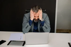 Man at a desk holding his head in his hands in front of a laptop